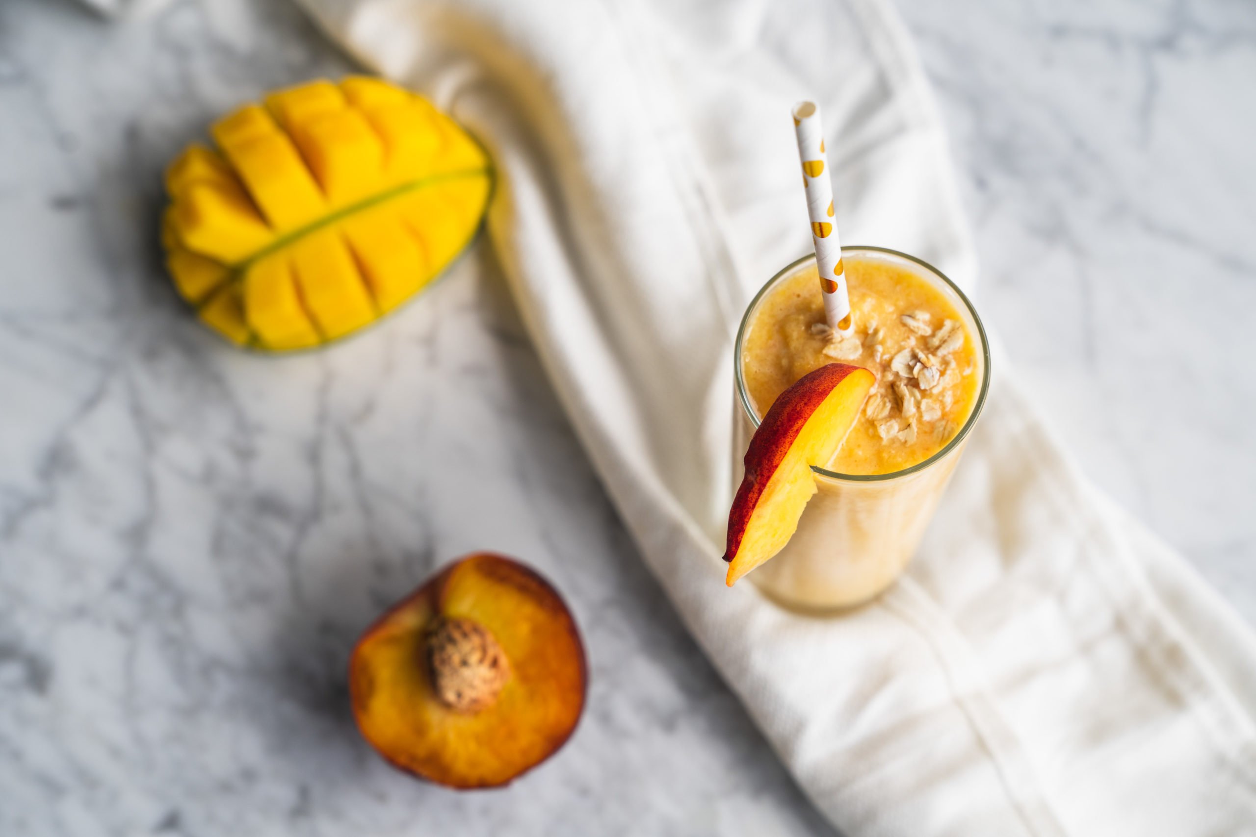 Delicious peach mango smoothie sitting on a table cloth with halves of mango and peach surrounding it.