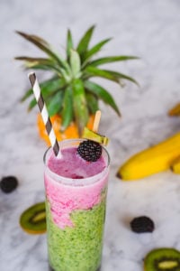 Kale smoothie with tropical berry top layer and surrounded by tasty fruit on a table
