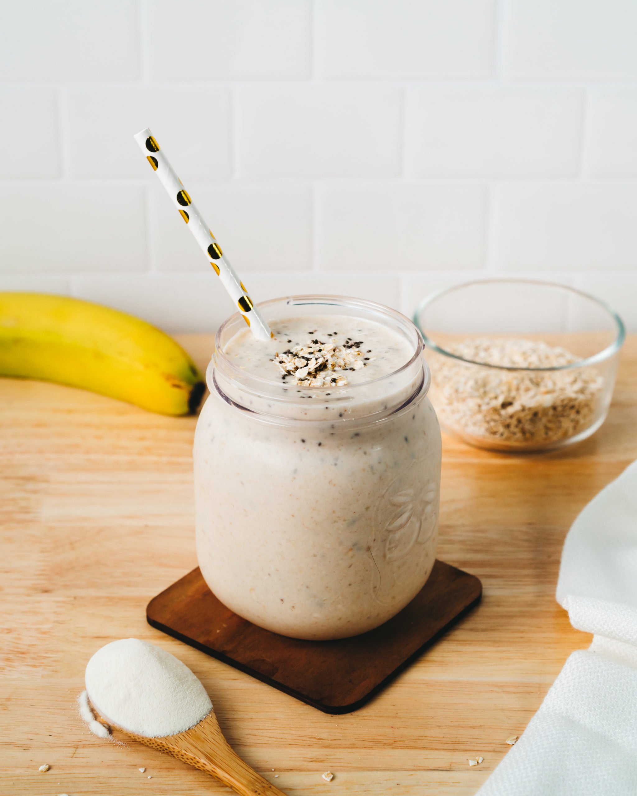 A delicious peanut butter banana smoothie in a mason jar with a straw, surrounded by ingredients including a table spoon of SkinnyFit Super Youth collagen