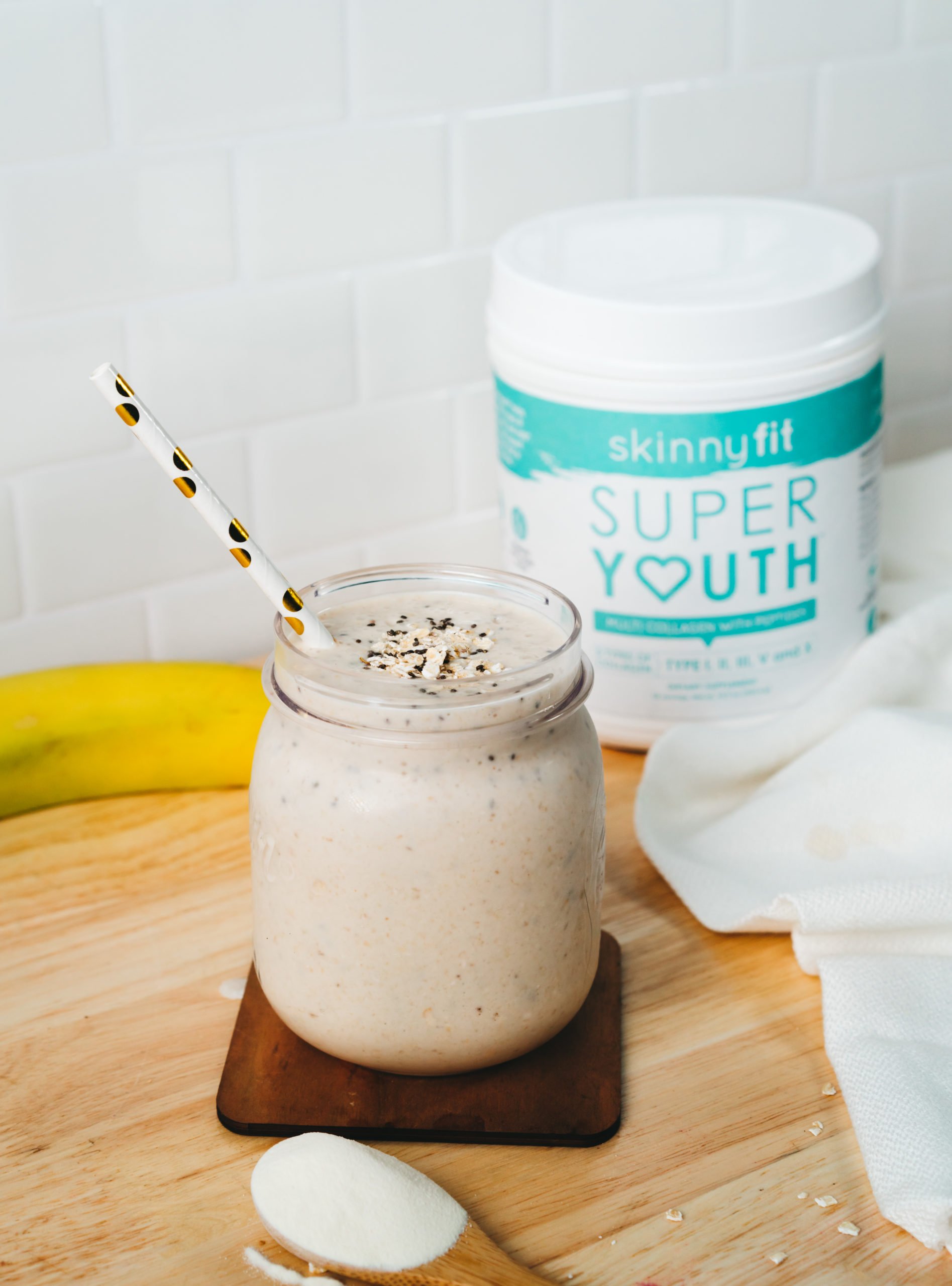 A mason jar filled with our peanut butter banana smoothie, with SkinnyFit Super Youth collagen, fruit, and other ingredients in the background