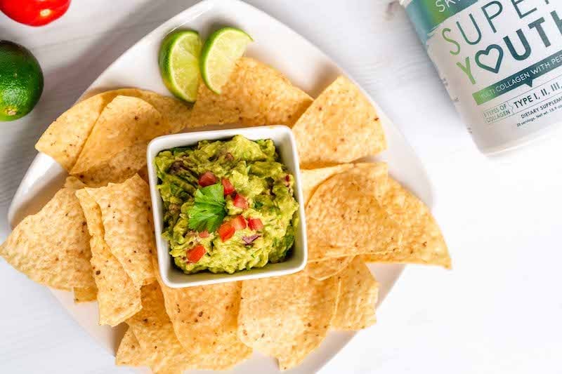 View of guacamole in serving bowel with chips, Super Youth collagen peptides and other ingredients needed to make our healthy guacamole recipe