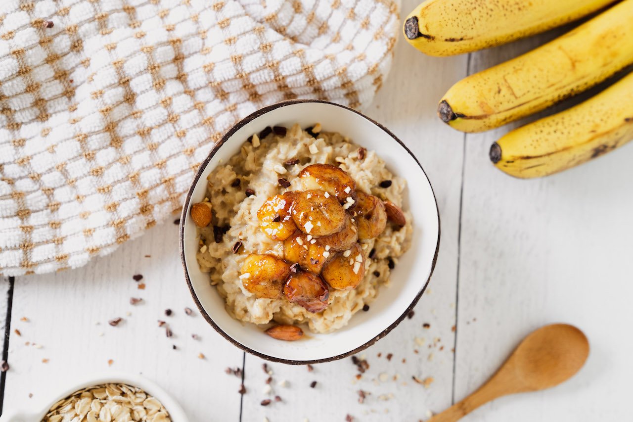Bananas Foster Oatmeal With Super Youth Collagen in a bowl surrounded by fresh bananas and cooking supplies.